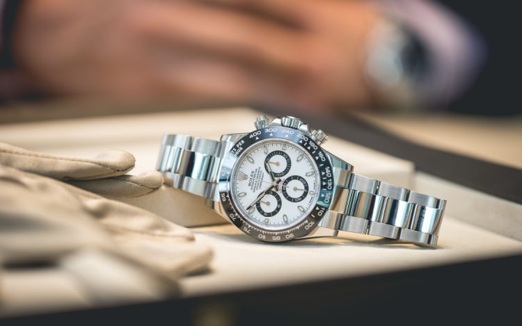A silver and white Rolex watch on a cream surface next to beige gloves, with a pair of hands blurred in the background