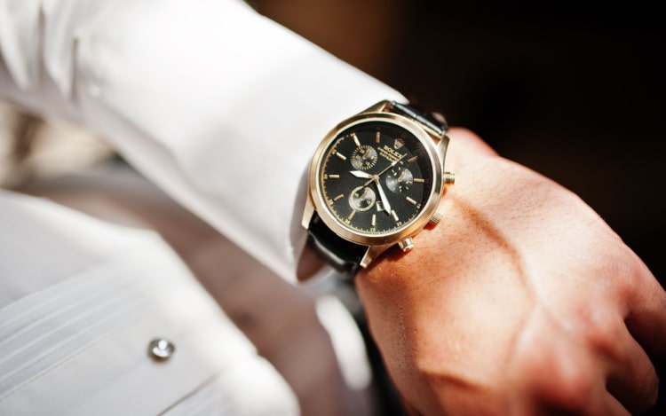a gold and black Rolex watch sits on a man’s wrist. He’s wearing a white shirt and the sun is shining on his hand, the watch, and his sleeve