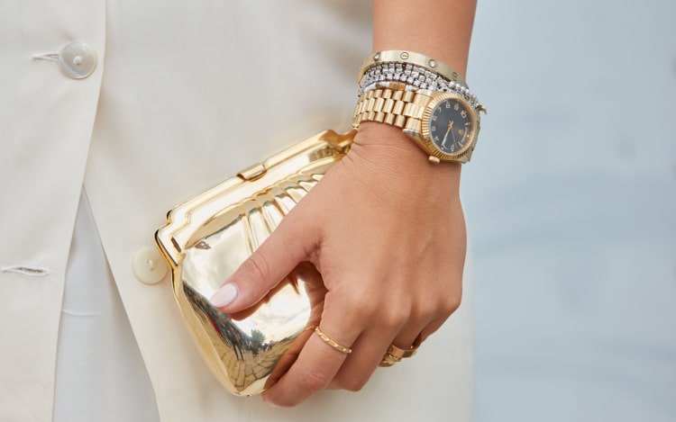 A gold Rolex watch with a dark face on a woman’s wrist next to a silver bracelet and a gold Cartier bangle.