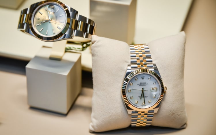 Two silver and gold Rolex watches. The watch on the left is on a cream-coloured stand and the watch on the right is wrapped around a cream-coloured cushion.
