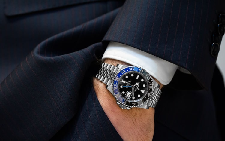 a silver Rolex with a black face on a man’s wrist. He’s wearing a navy suit and his hand is in his pants pocket