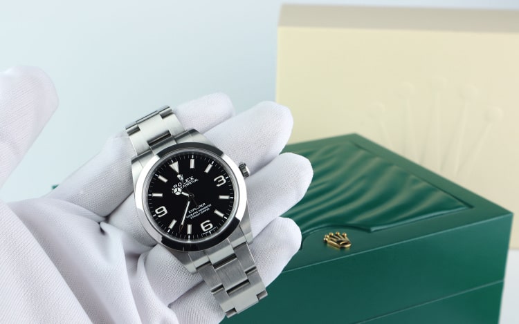 A silver Rolex watch being held by a white-gloved hand. A green watch box with a gold Rolex logo is in the background
