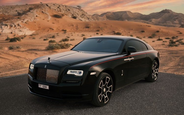 A black Rolls-Royce car, stationary on a dark road with the dessert behind it.