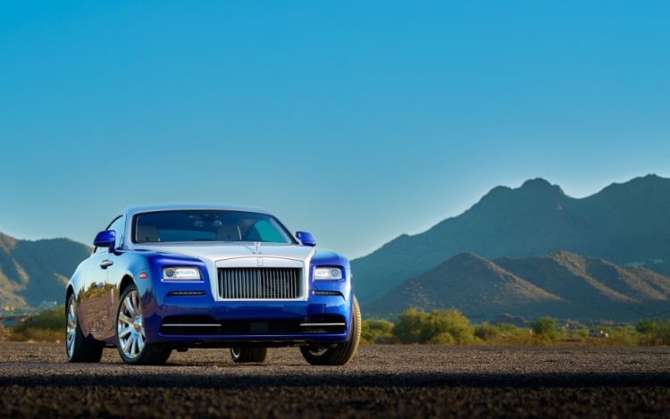 A dark blue Rolls-Royce car stationary on a dirt road with mountains and blue skies behind it.