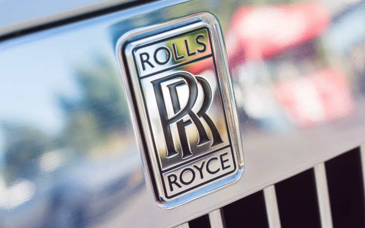 A close-up of a silver Rolls-Royce car showing the emblem, two R’s overlapping each other with the word ‘Rolls’ above and the word ‘Royce’ below.