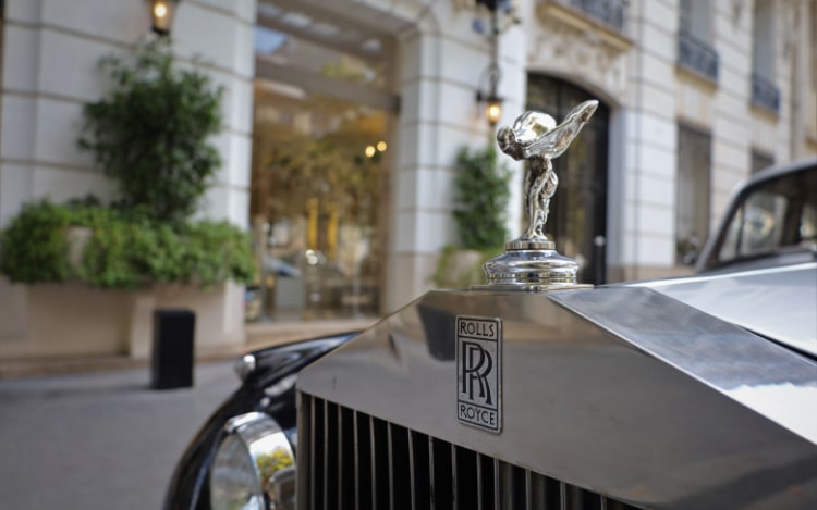 A close-up of a silver Rolls-Royce showing the Spirit of Ecstasy hood ornament, a silver winged woman leaning forward.