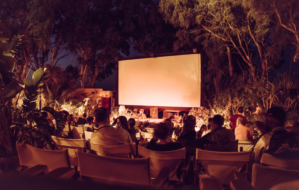 Kamari town open air cinema. People sat on chairs facing a projector waiting for the movie to begin with no roof and surrounded by trees