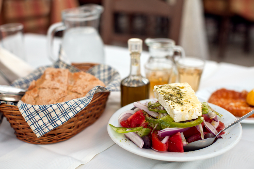 A white table with a plate of greek salad, a basket of bread and bottles of liquid blurred behind them
