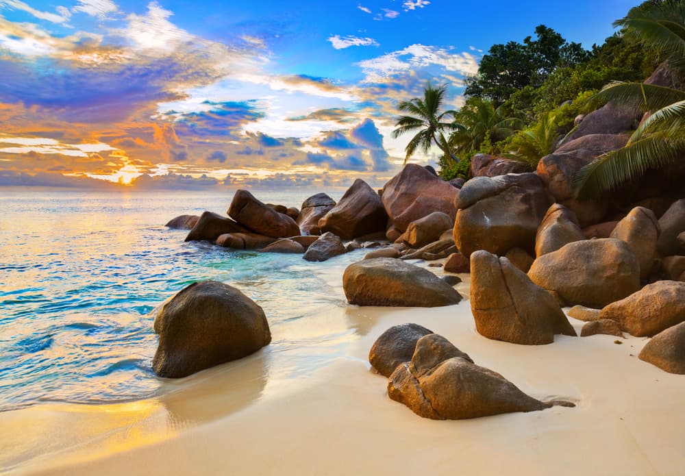 Tropical beach sunset with blue water, a brown pile of rocks and green palm trees in the upper right corner