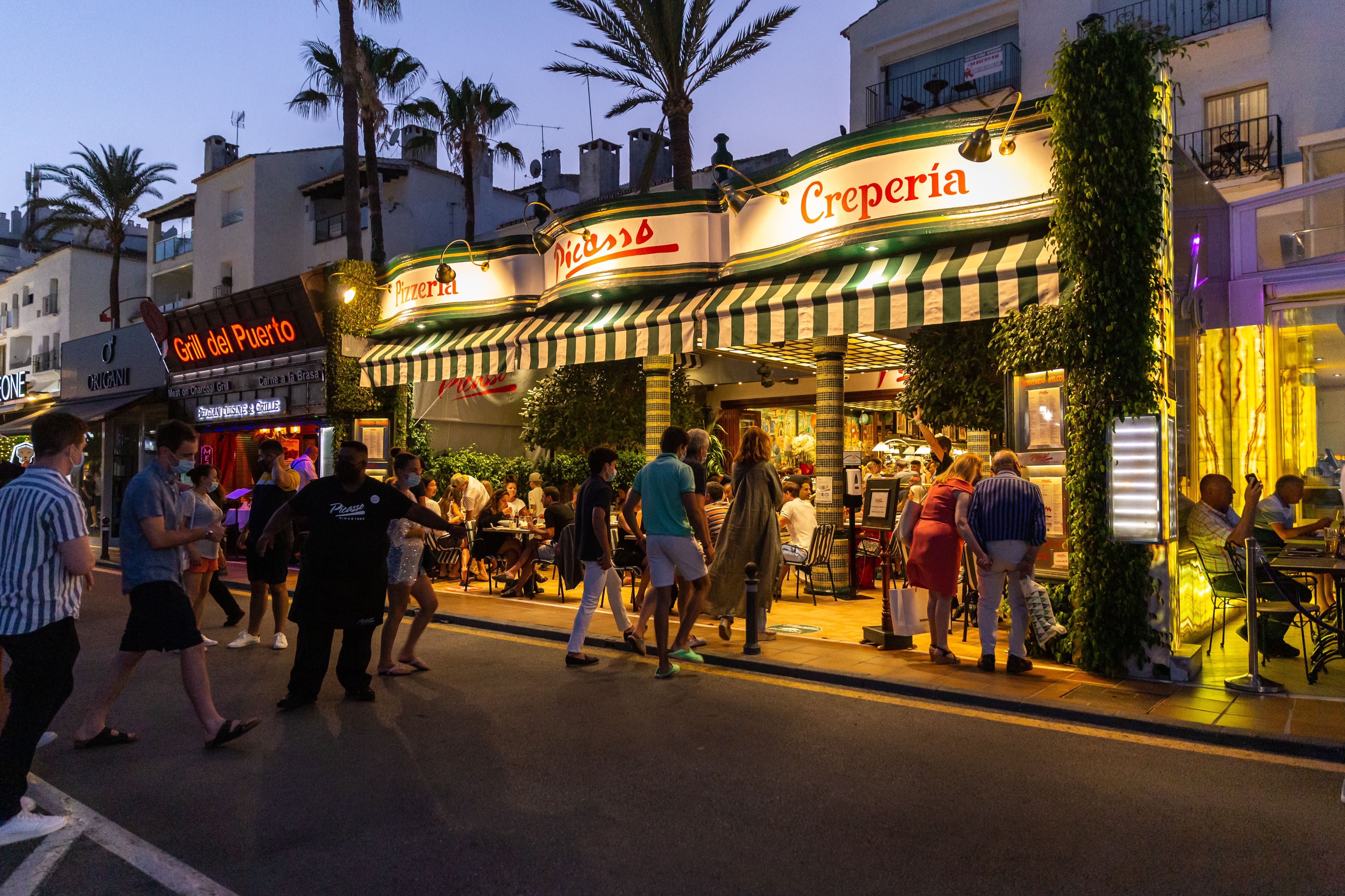 Busy Spanish street at nighttime with a queue of people walking into a well-lit and busy restaurant.
