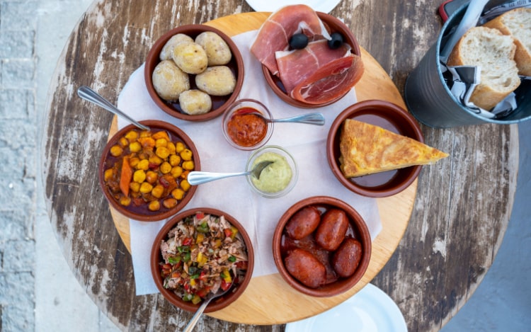 Multiple tapas in terracotta bowls with serving spoons, including chorizo, a slice of Spanish tortilla, cured ham, a chickpea dish, bread, and two condiments in clear glass dishes.