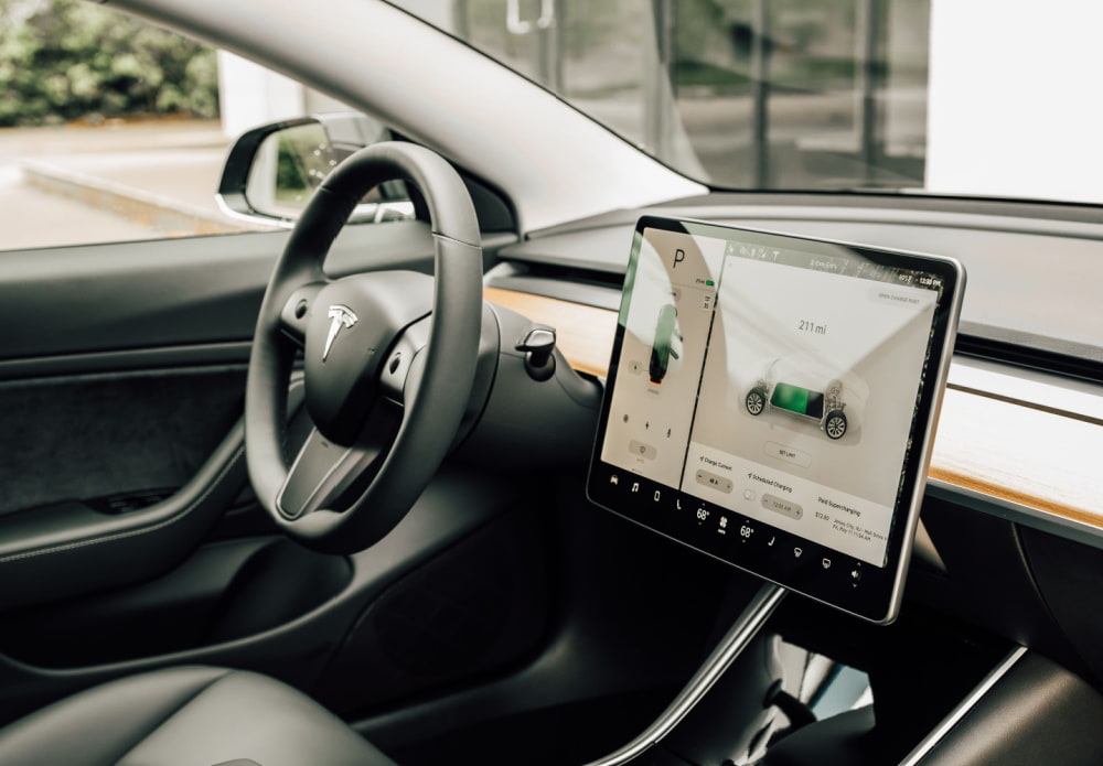Side shot inside a car showing the steering wheel and the end of the driver's seat. To the right of the steering wheel is a large screen/control centre with details of the car pictured on it.