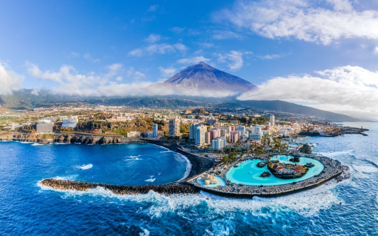 Tenerife’s coast taken from the ocean, with dark blue water, a bright blue pool, a rocky coastline, scattered buildings and a mountain partially covered in cloud