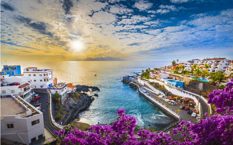 Tenerife looking out to the sea where the sun is shining yellow and the sky is covered in wispy clouds with the blue sky visible behind them.