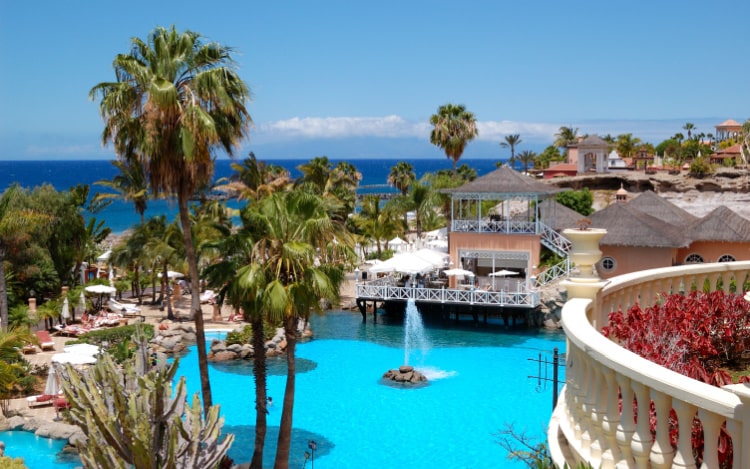 A luxury resort in Tenerife with palm trees, a bright blue pool, a terrace with seating and white umbrellas, and a white balcony with dark pink flowers