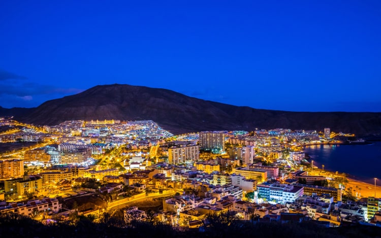 enerife at night under a dark blue sky with a mountain barely visible in the distance. The buildings are all illuminated with warm light