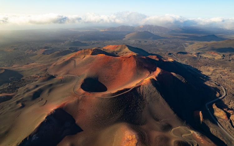 An aerial view of a dormant volcano in Timanfaya National Park. The volcano is mostly dark grey in colour with large flecks of sandy red.