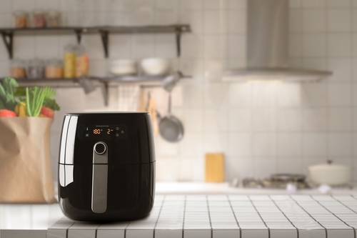 Black air fryer photographed in a modern and minimalistic chrome and white kitchen. Sat on a checkered work surface with a brown paper bag of vegetables to the left and a stovetop to the right.
