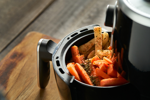 Black air fryer open slightly with seasoned carrots and potatoes in, set on a wooden work surface.