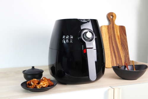 Black and silver air fryer on a wooden work surface with a plate of crisp octopus and seasoning dish to the left and an array of serving spoons and a wooden chopping board to the right.