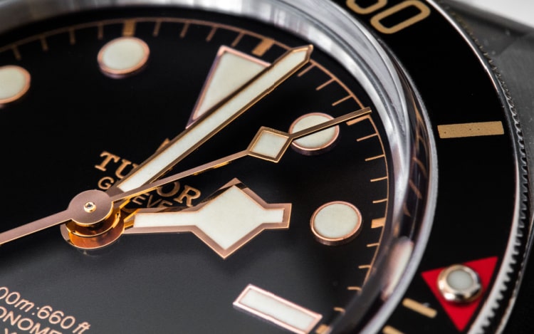 A close-up image or a black Tudor watch with white and gold detailing on the face and dial.