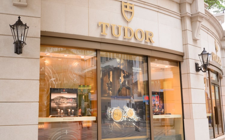 A Tudor storefront. The building is light coloured stone with the Tudor logo above the windows in gold. In the windows are 3 Tudor posters and several watches.