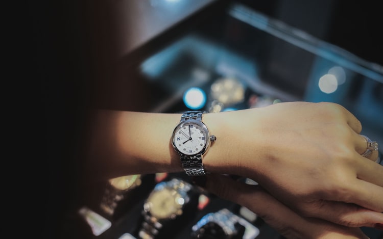 A silver Tudor watch with a white face on a woman’s left wrist. In the background, slightly out of focus, is a glass cabinet with more watches.