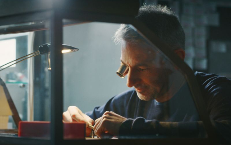 A grey-haired male watchmaker sat at his workbench with an eyeglass in his right eye, peering down at the watch he is making.