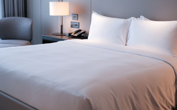 A freshly made bed with white pillows and a white duvet cover. In the left hand corner is a bedside table with a phone and a lamp on top