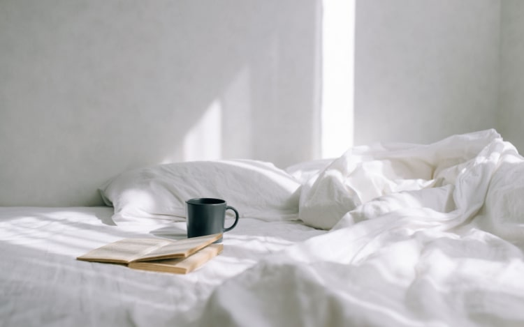A grey coffee mug and an open paperback book sit on an unmade bed with white pillows, a white fitted sheet, and white duvet cover.