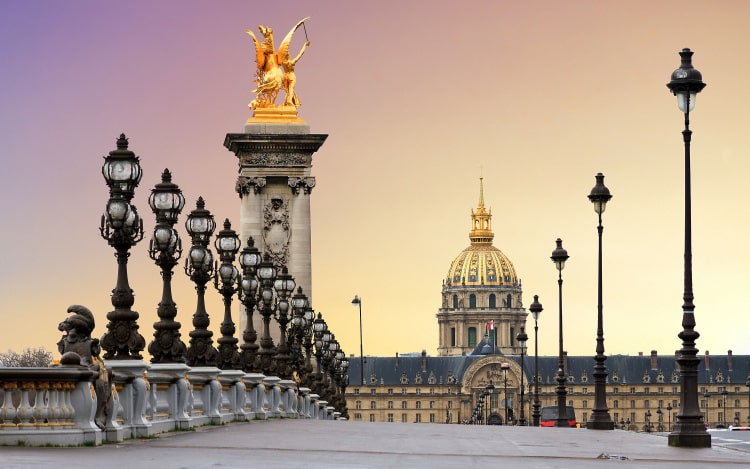 A view over a bridge in Paris, with several black lampposts lining the bridge, an ornate gold statue on a plinth, and a large building in the background.