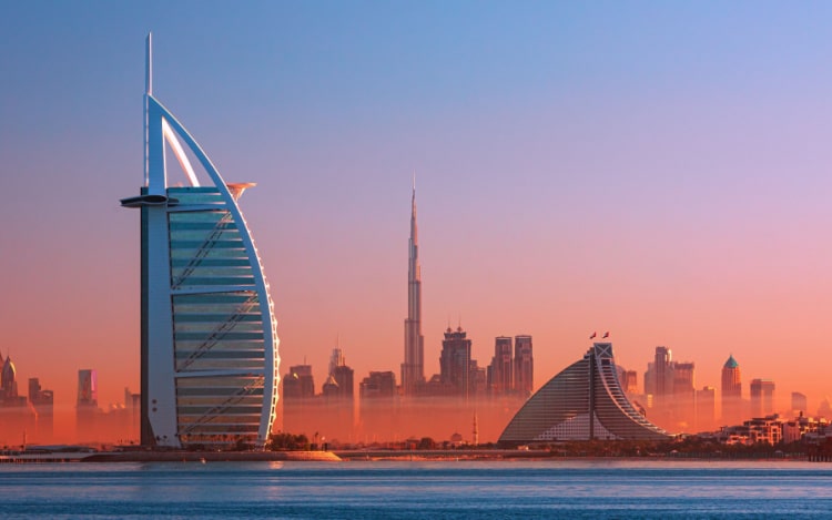 The Burj Al Arab at sunset, with other skyscrapers in the distance under a blue, pink and orange sky.