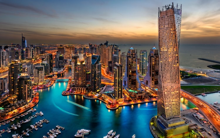 Dubai skyline at dusk, with several skyscrapers illuminated and a view of a port with multiple boats.