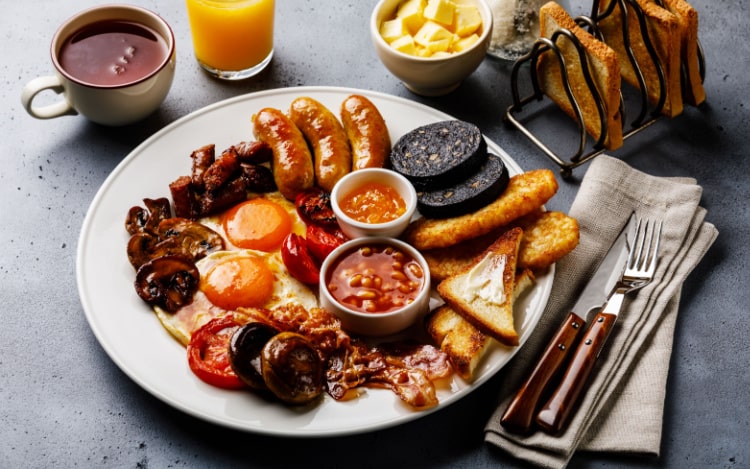 A full English breakfast on a white plate, a toast rack, a small bowl of butter, a cup of tea and orange juice, and cutlery sit on a grey table.