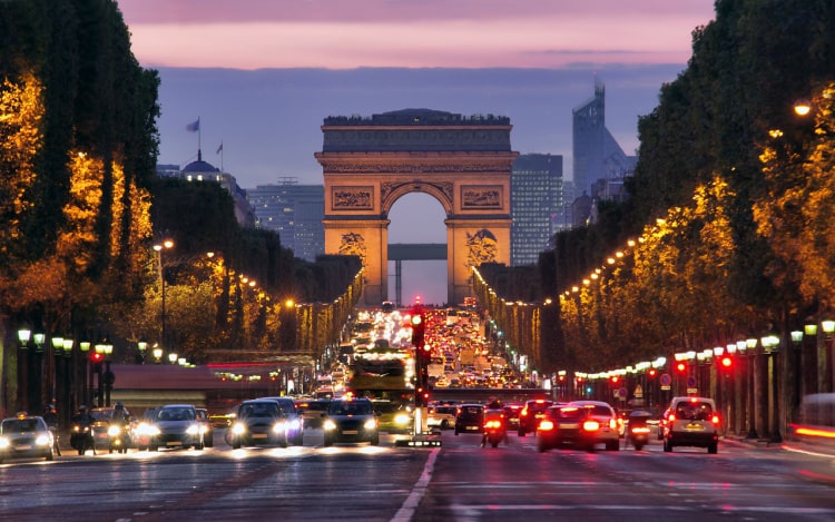 A main road in Paris at dusk with several cars, trees lining the street, and the Arc de Triomphe monument in the centre.