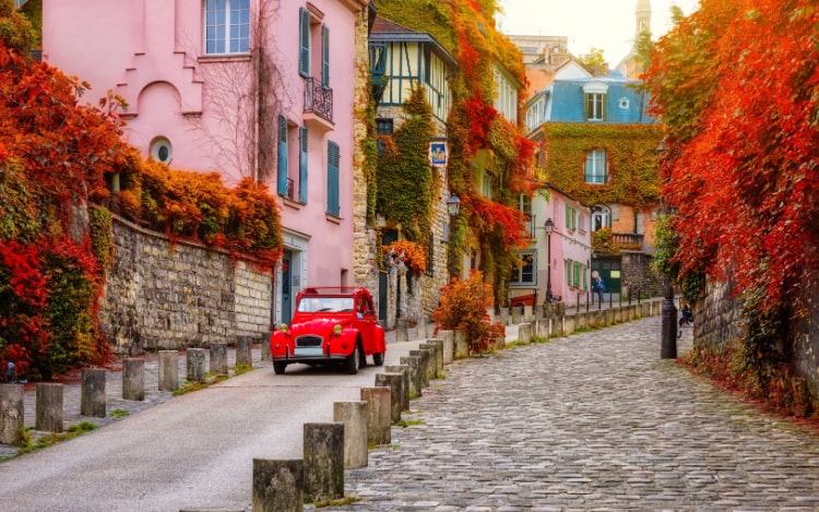 A cobblestone street in the Montmartre area of Paris. A bright red car is parked next to a pink house and the leaves on the trees and bushes are shades of red and orange.