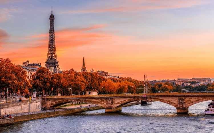 A view of Paris from the Seine. The Eiffel Tower is on the left, with several trees and a bridge running across the water. The sky is shades of orange, pink and blue.