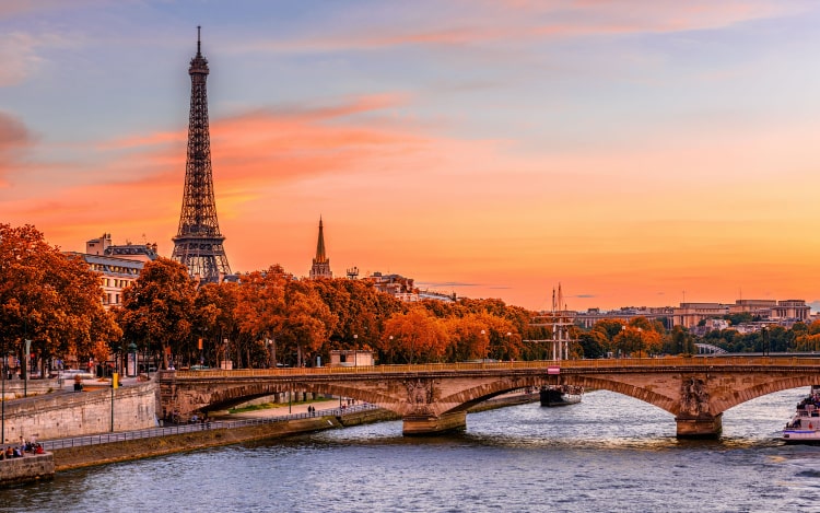 A view of Paris from the Seine. The Eiffel Tower is on the left, with several trees and a bridge running across the water. The sky is shades of orange, pink and blue.