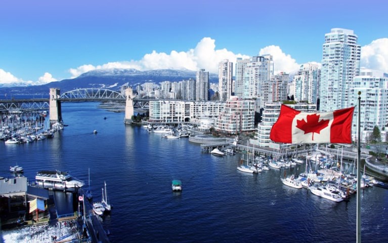 A coastal city in Canada on a clear day, with the Canadian flag flying to the right along with light-coloured skyscrapers.