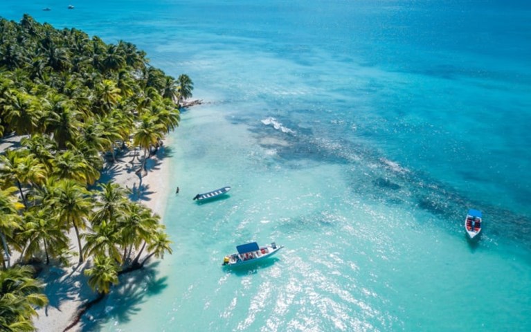 aerial view of an island’s coast, with turquoise blue waters, two boats floating and palm trees on a beach