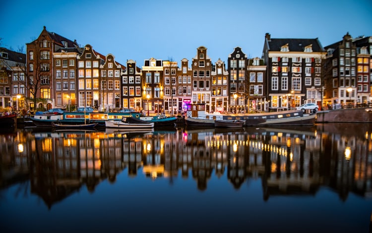 A row of houses in Amsterdam in the evening, reflecting in the still waters of the canal with a few stationary boats.