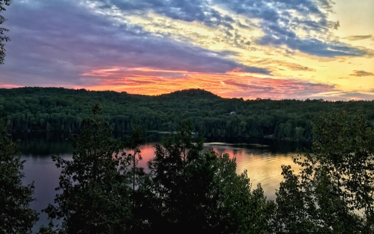 A lake at sunset surrounded by green trees under a yellow, blue and pink sky.