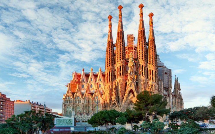 La Sagrada Familia partially lit up by the setting sun under a blue and cloudy sky with trees at the bottom of the image.