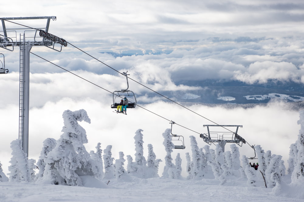 Two people going up a snowy mountain on a chairlift with snowy trees and clouds in the background