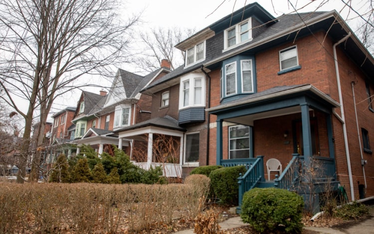 A row of a few houses in a suburban neighbourhood. The focal point is a red brick house with blue painted window frames and a blue porch.