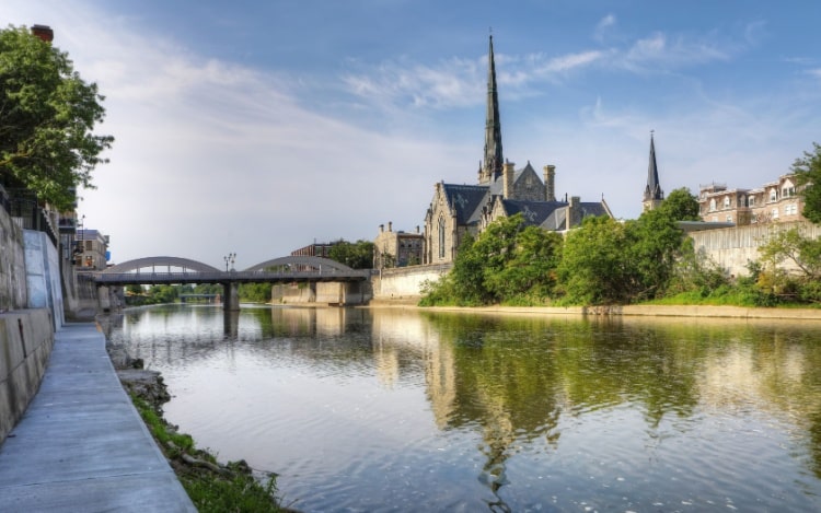 A path next to a large river on a clear blue day with a bridge in the distance and a church and houses visible on the other side of the river.