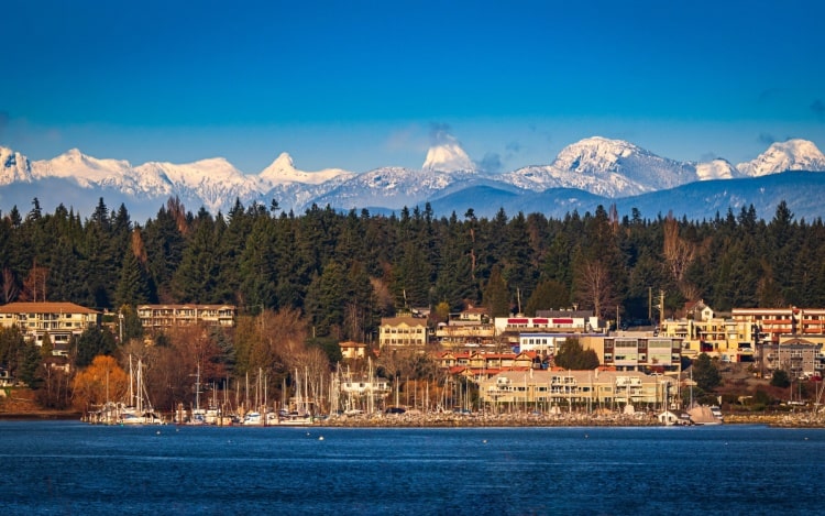 A view of Comox Valley from the water, with buildings and boats on the shoreline, trees behind and snow-capped mountains in the background.