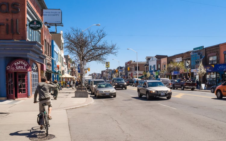 A main street in Danforth, with multiple cars on the road and a cyclist riding away from the image, next to a building with a red door that says ‘Pappas Grill’.