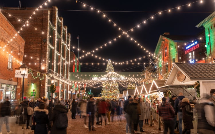 Distillery Winter Village Christmas market at night, with rows of fairy lights, several people, and a lit-up Christmas tree in the background.