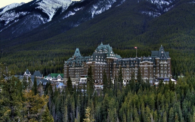 The Fairmont Banff Springs hotel, a large brown castle-like building, is nestled between hundreds of tall green trees and snow-capped mountains.
